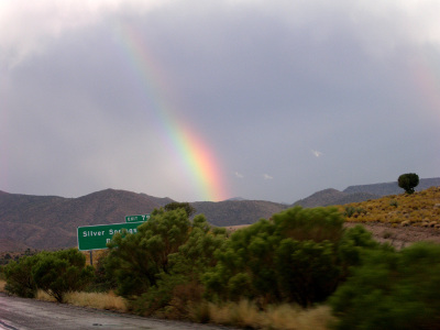 [Seeming to start behind the mountains in the distance, a colorful stripe extends skyward at a leftward slant. The lower part of the rainbow is brighter and all the colors more visible with violet on the left and red on the right with green, yellow, and orange in between. The upper part of the rainbow seems to run up through some dark clouds which mute the color in the upper part of the stripe. There is a roadsign with an exit for Silver Springs behind some large green shrubbery.]